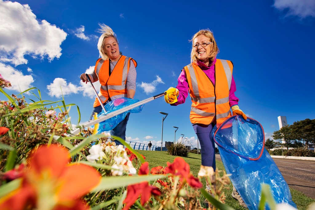 Two women with blond hair wearing hi-vis jackets. They are carrying litter pickers and blue bin bags, and collecting litter from a flowerbed. 